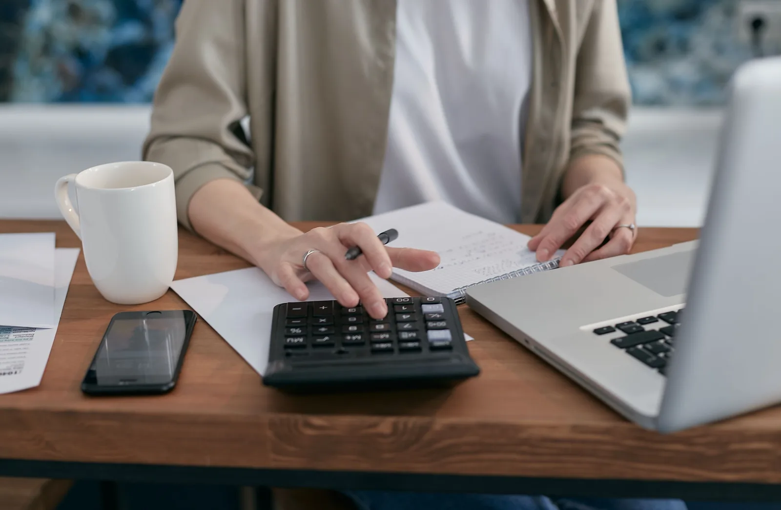 A person using a calculator and laptop while taking notes at a wooden desk. 1099 vs W-2 for Bookkeeping Firm Staff: The Real Test